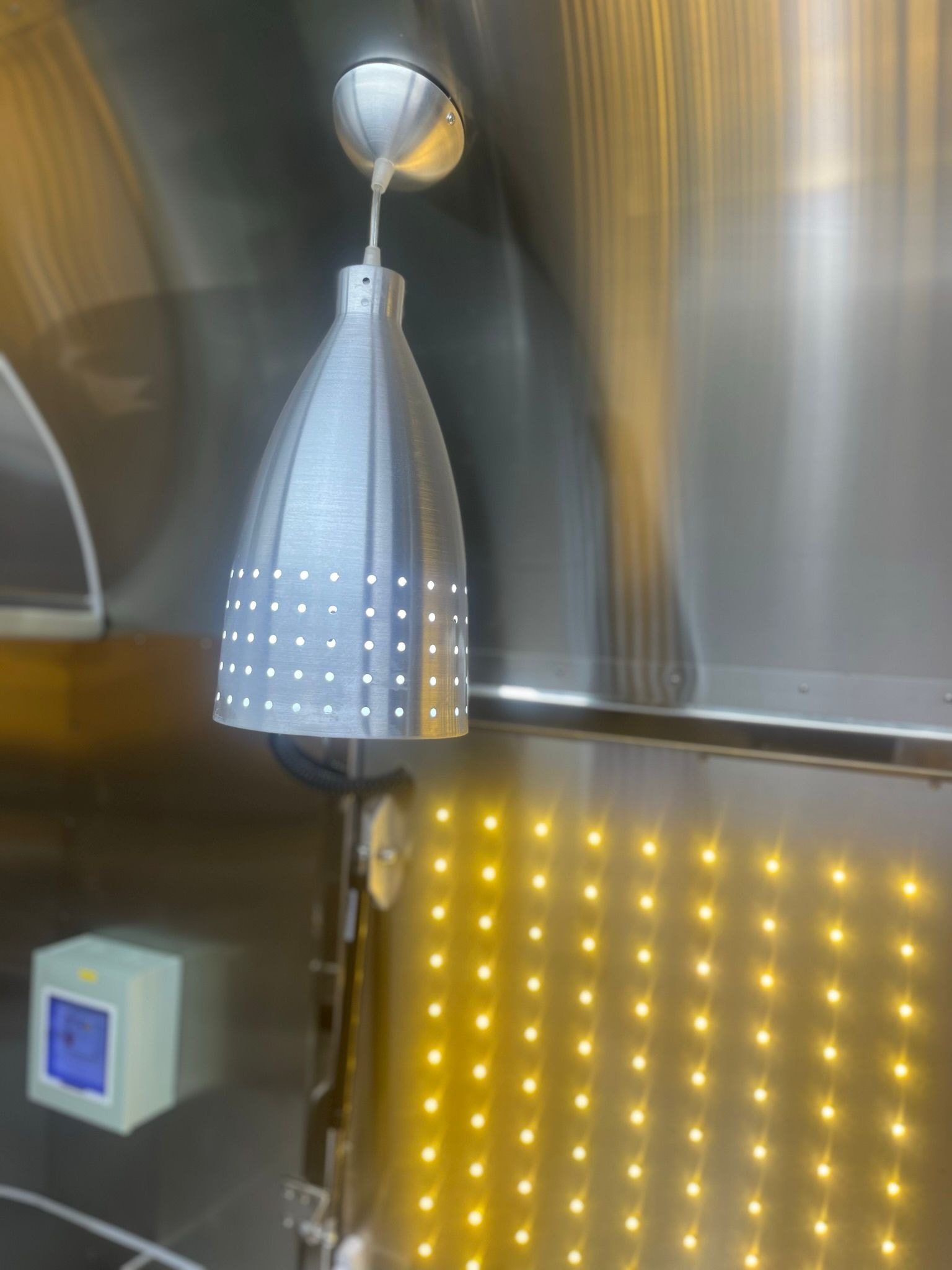 Brushed aluminium perforated pendant light inside Airstream trailer with warm LED display panel behind